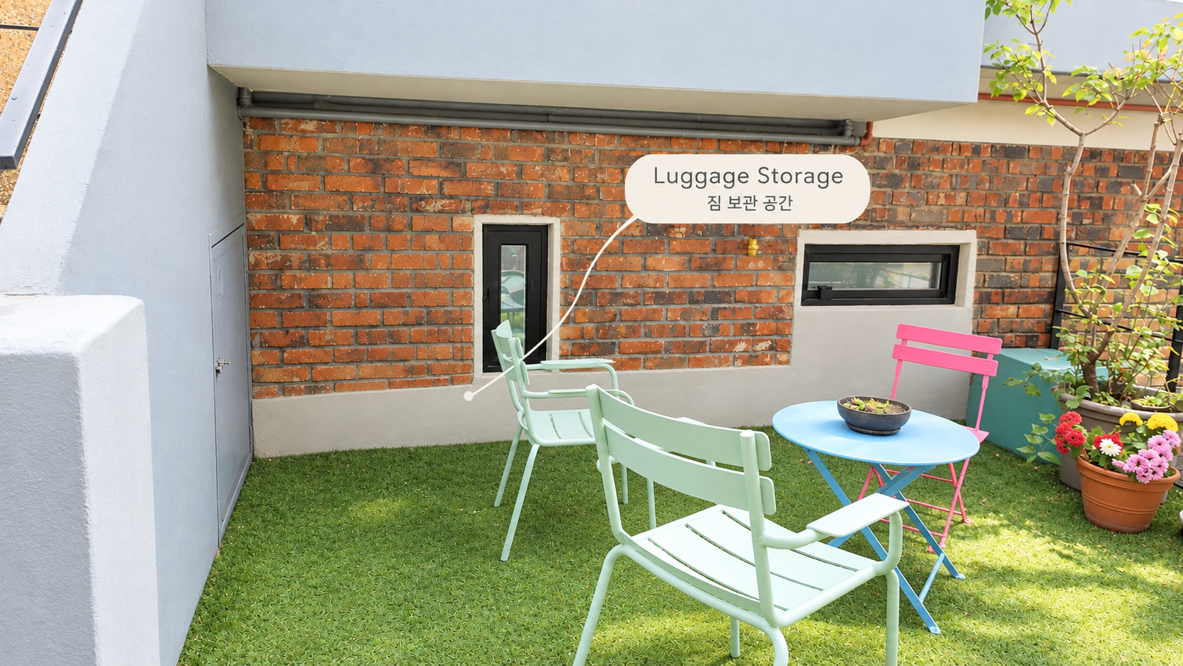 The ground-floor garden with artificial grass, mint and pink chairs and a blue table. A label marks the luggage storage spot under the covered overhang beside the brick wall.